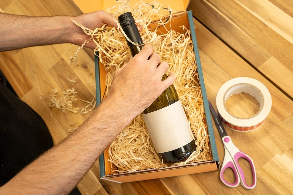 Hands packing a bottle of white wine into a shipping box filled with wood wool